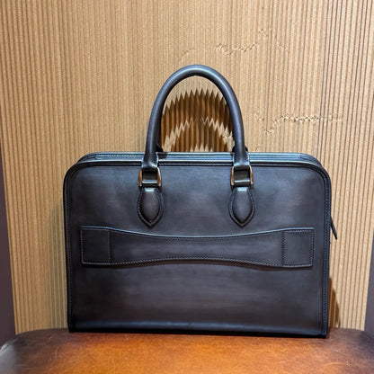 A dark brown leather briefcase with double top handles and brass hardware, displayed on a wooden surface against a corrugated cardboard background.