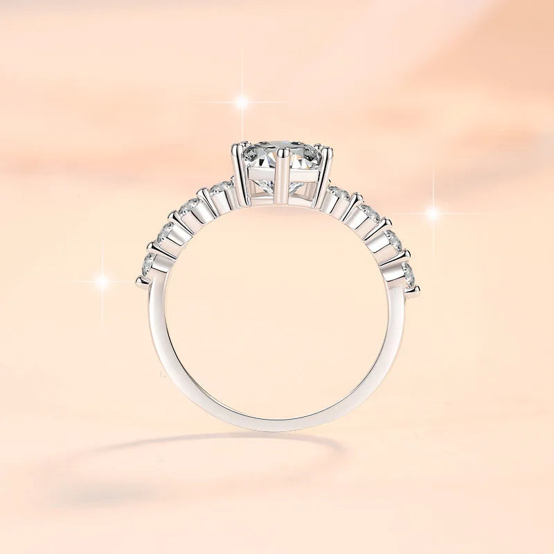 A silver-toned diamond ring with a large round center stone and smaller diamonds along the band, shown against a soft peach background with sparkling light effects.