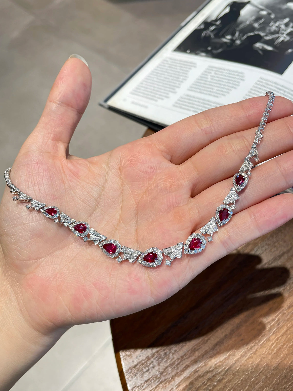 A silver-toned diamond necklace with red gemstone accents is displayed on an open hand. The necklace features multiple oval-cut red gemstones surrounded by sparkling diamond settings.
