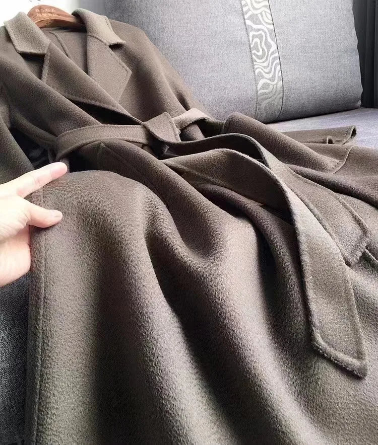 A close-up of a taupe-brown wool coat draped over a gray sofa, with a hand holding the fabric to show its soft texture.