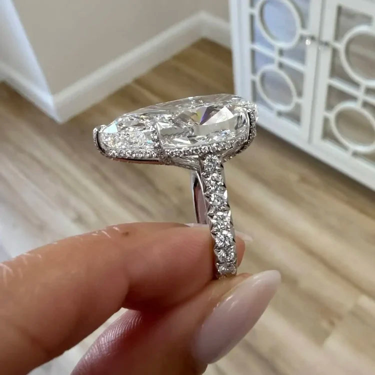 A close-up of a hand holding a silver engagement ring with a large marquise-cut clear gemstone and a pavé-set band, with a blurred indoor room in the background.