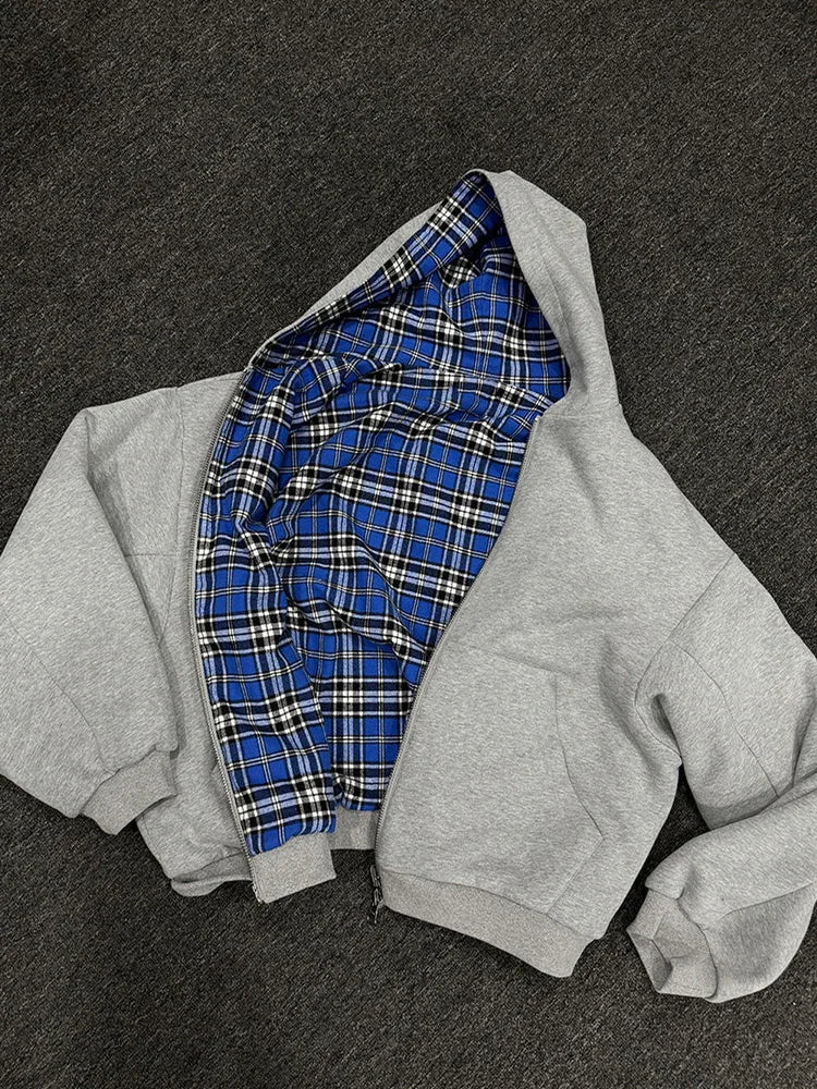 A light gray zip-up hoodie laid flat on a dark gray textured surface, with the hood open to reveal a blue, black, and white plaid lining.