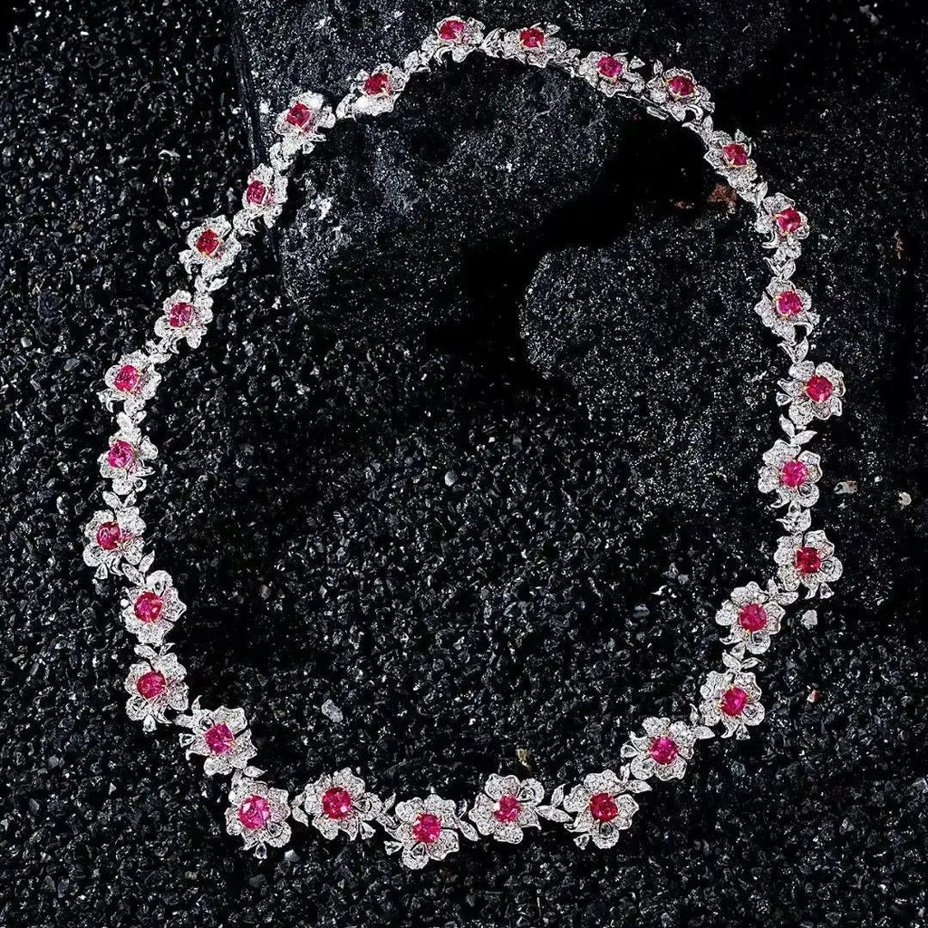 A floral necklace featuring silver-toned settings with small clear stones and bright pink gemstones, arranged in an oval on a dark textured surface.