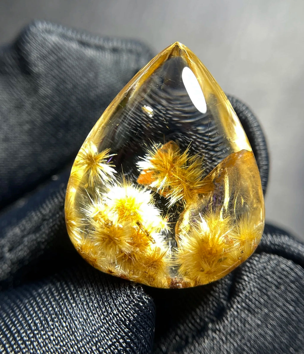 A teardrop-shaped clear resin cabochon containing golden-yellow dried flower buds, resting on dark textured fabric.
