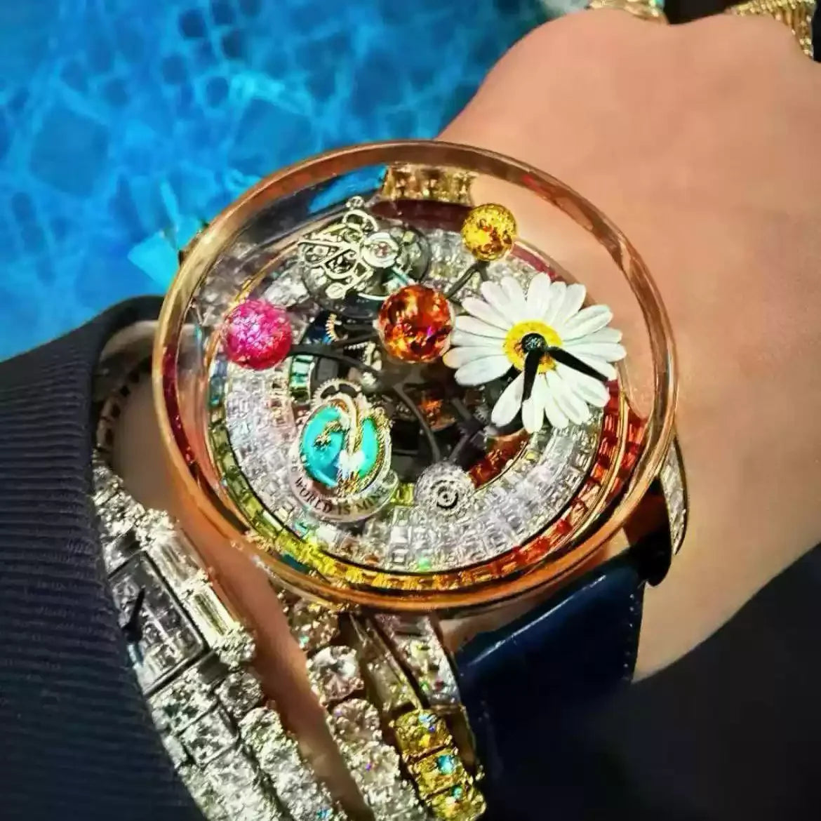 A close-up lifestyle shot of a heavily embellished luxury watch on a person's wrist. The watch features a rose gold case, a clear crystal dome filled with intricate mechanical gears, colorful gemstones, a white daisy flower, and a dark blue leather strap.