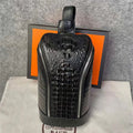 A black crocodile-embossed leather backpack with a top handle, displayed on a light beige dust bag in front of its orange and black packaging box.