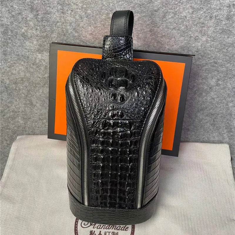 A black crocodile-embossed leather backpack with a top handle, displayed on a light beige dust bag in front of its orange and black packaging box.