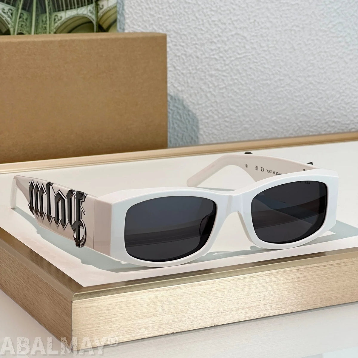 White rectangular sunglasses with dark lenses and a black Gothic-style 'malay' graphic on the temple arm, displayed on a light wood tray.