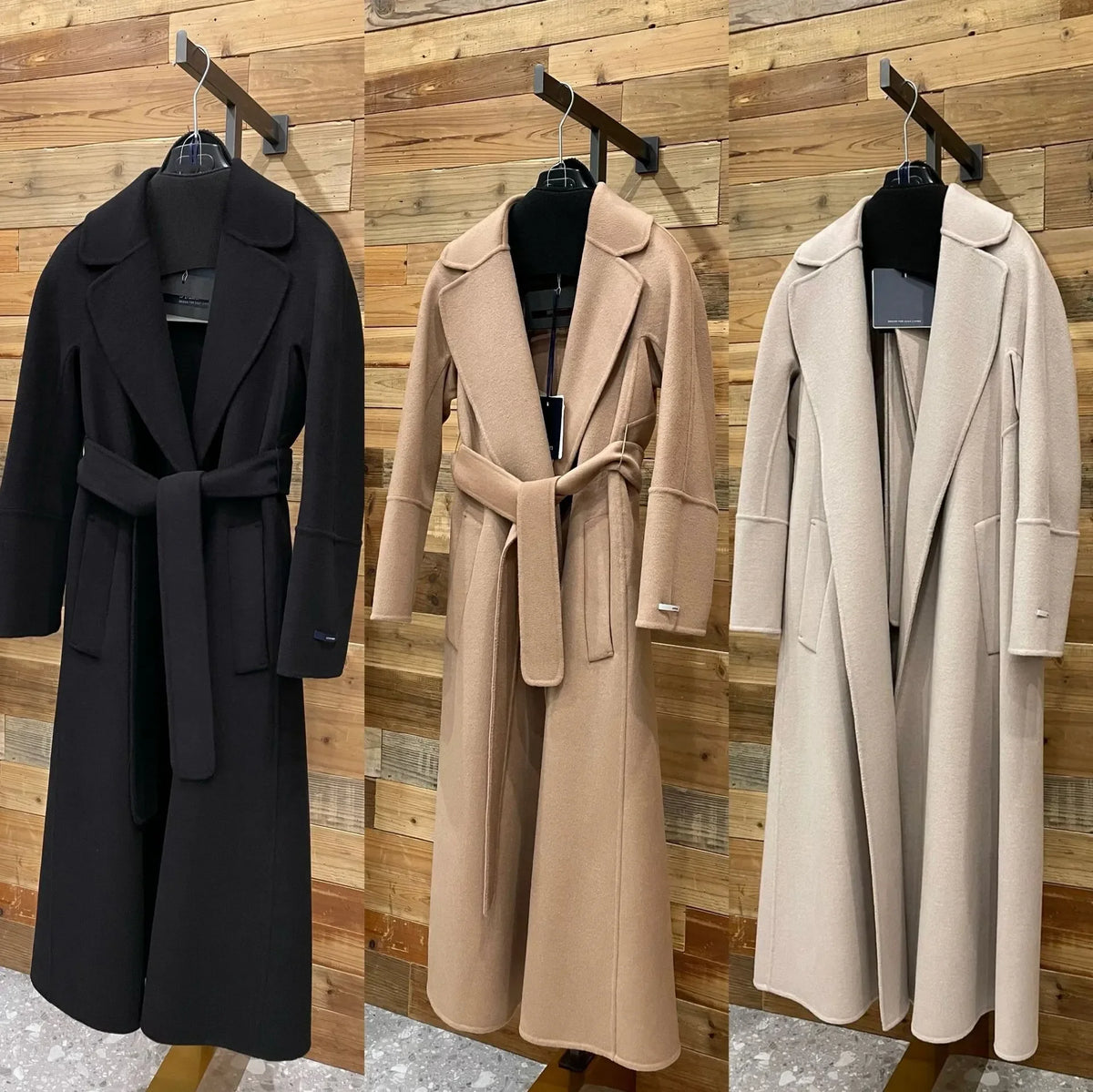 Three long belted wool coats displayed side by side on black hangers against a wood plank wall. The coat on the left is black, the middle is camel beige, and the right is light beige/cream.