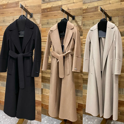 Three long belted wool coats displayed side by side on black hangers against a wood plank wall. The coat on the left is black, the middle is camel beige, and the right is light beige/cream.