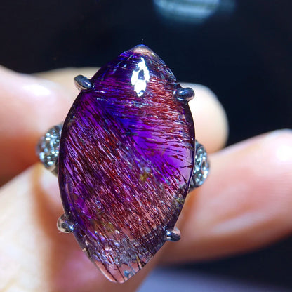 A close-up of a ring held between fingers, featuring a large marquise-shaped purple gemstone with fine metallic needle-like inclusions and silver-toned prongs and band details.
