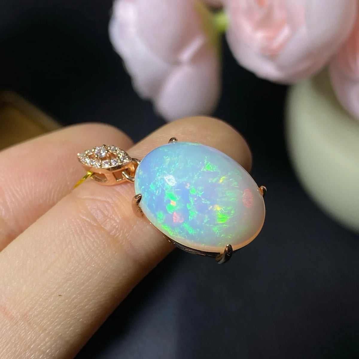 A close-up of a hand holding a rose gold ring with an oval white opal cabochon and a small diamond accent on the band, with soft pink flowers blurred in the background.