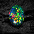A vibrant, oval-shaped opal gemstone displaying a multicolor play-of-color with bright green, blue, red, and yellow flashes, set against a dark textured wood background.