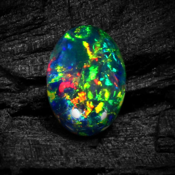 A vibrant, oval-shaped opal gemstone displaying a multicolor play-of-color with bright green, blue, red, and yellow flashes, set against a dark textured wood background.