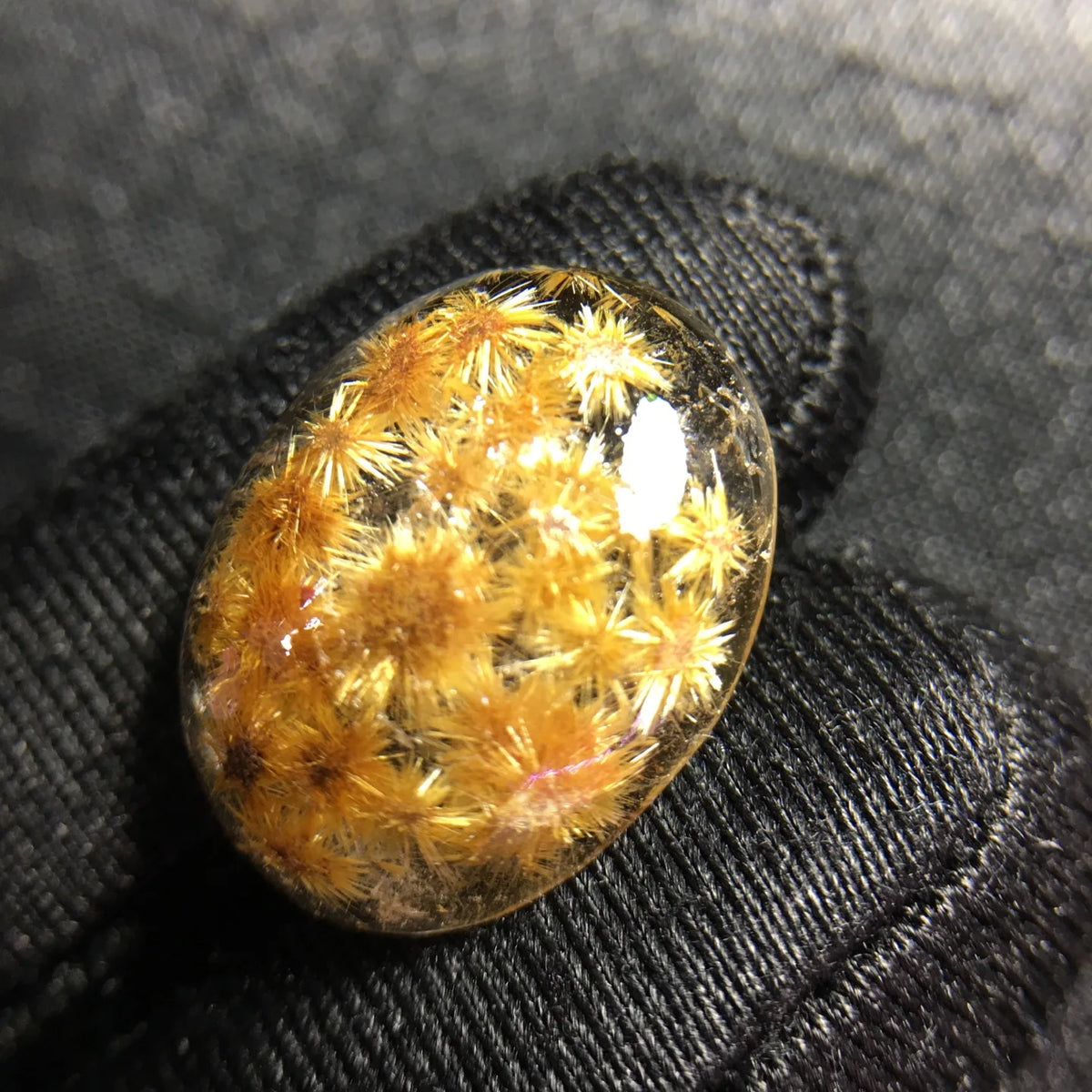 A polished oval gemstone containing golden-yellow crystalline structures, resting on a black textured fabric surface.