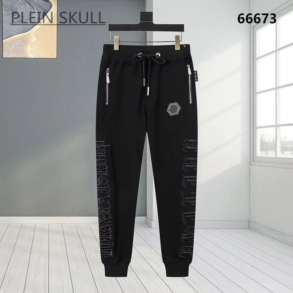 Black jogger pants with silver zipper details, a drawstring waist, and vertical gothic lettering on the legs, shown hanging against a modern gray and white interior background.
