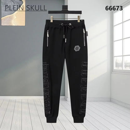 Black jogger pants with silver zipper details, a drawstring waist, and vertical gothic lettering on the legs, shown hanging against a modern gray and white interior background.