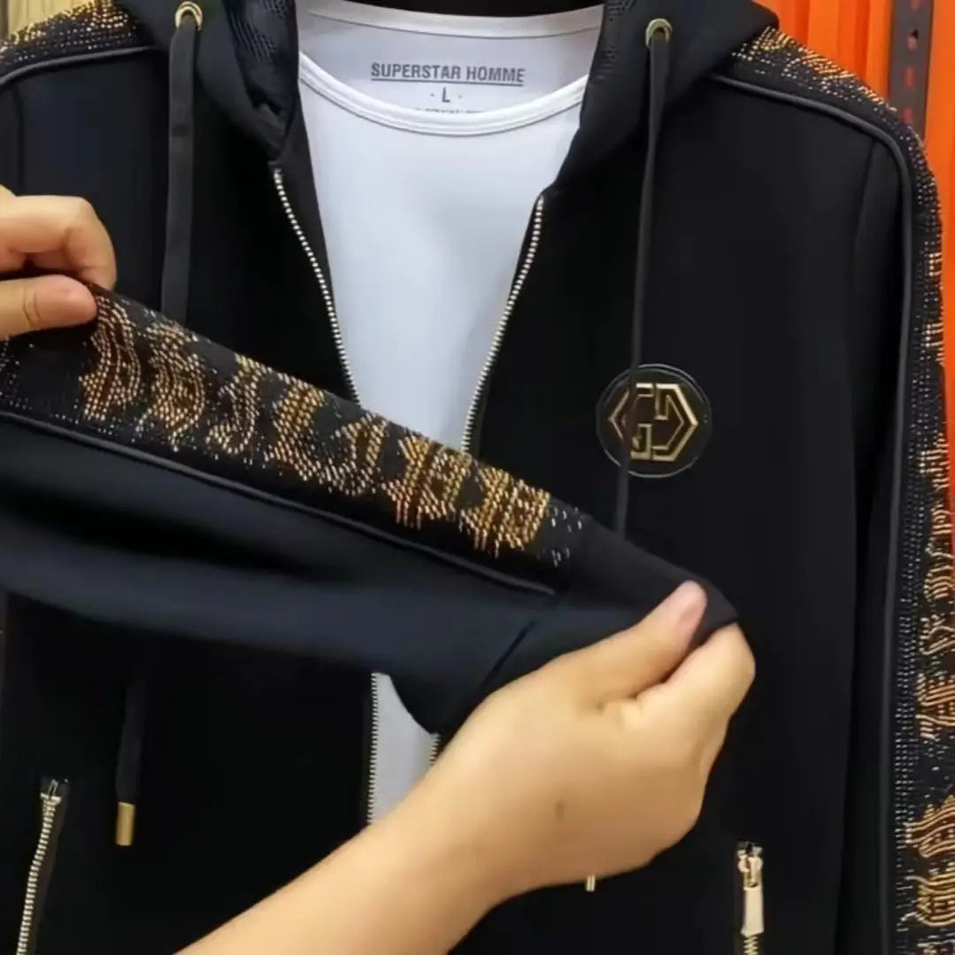 A black zip-up hoodie with gold rhinestone-embellished sleeve panels and a gold circular chest emblem, shown being held open by hands over a white t-shirt.
