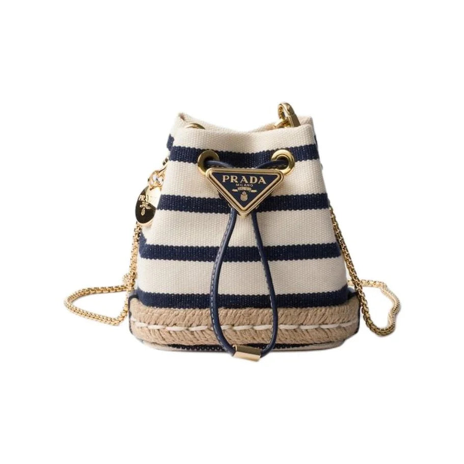Prada striped canvas bucket bag with navy blue and cream horizontal stripes, a gold-tone triangular Prada logo plaque, a woven jute base, and a gold chain strap, shown on a plain white background.