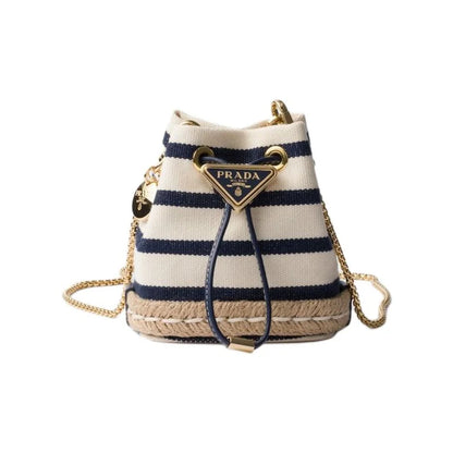 Prada striped canvas bucket bag with navy blue and cream horizontal stripes, a gold-tone triangular Prada logo plaque, a woven jute base, and a gold chain strap, shown on a plain white background.