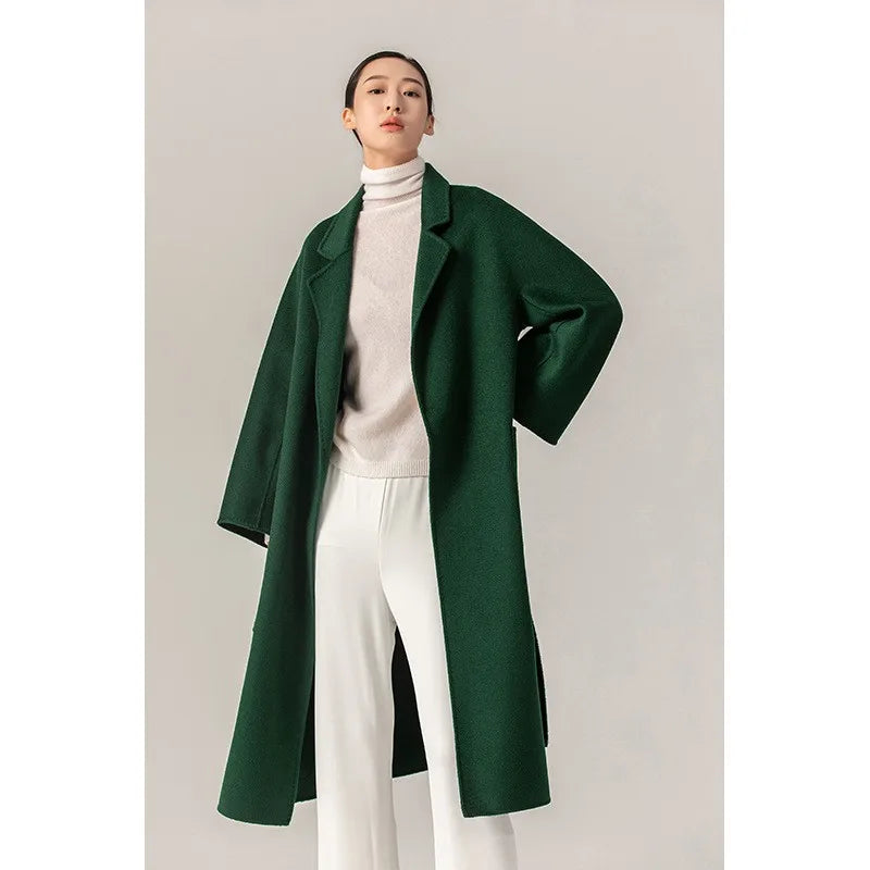 A woman modeling a long dark green wool-blend coat over a cream turtleneck sweater and white wide-leg trousers, standing against a plain light gray studio background.