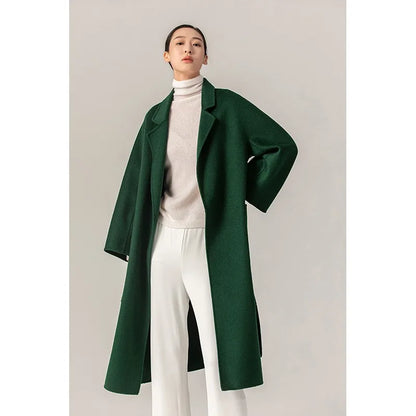 A woman modeling a long dark green wool-blend coat over a cream turtleneck sweater and white wide-leg trousers, standing against a plain light gray studio background.