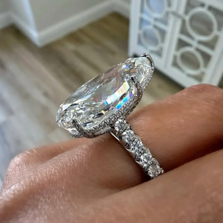 A close-up of a hand wearing a large oval clear gemstone ring with a silver-toned pavé band, photographed indoors with a blurred background.