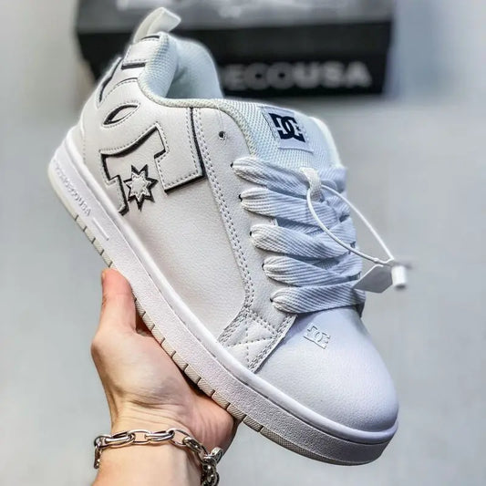 A hand holds a white DC Shoes sneaker with black embroidered details and white laces, with a blurred DC box in the background.