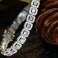 A close-up of a diamond bracelet featuring rectangular baguette-cut clear stones in silver-toned settings, arranged diagonally against a dark brown textured background.