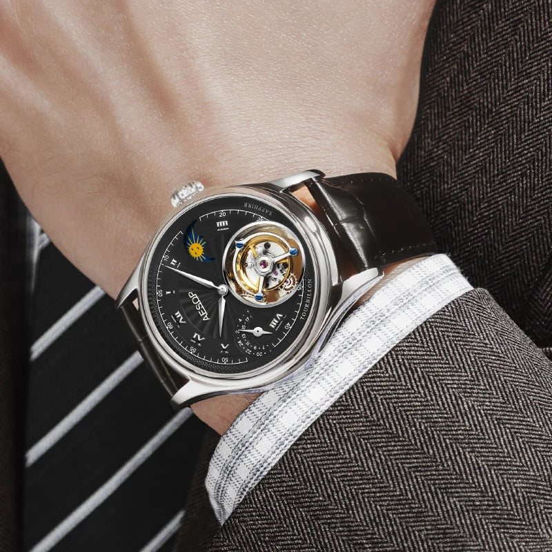 A close-up of a wrist wearing a luxury watch with a black dial, silver case, and black leather strap, styled with a striped tie and textured gray suit jacket.
