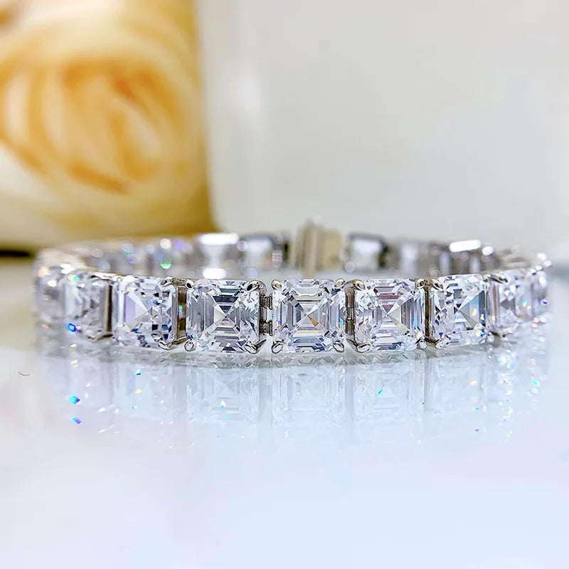 A silver-toned tennis bracelet featuring a row of square-cut clear gemstones, displayed on a reflective white surface with a soft-focus beige floral background.