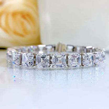 A silver-toned tennis bracelet featuring a row of square-cut clear gemstones, displayed on a reflective white surface with a soft-focus beige floral background.