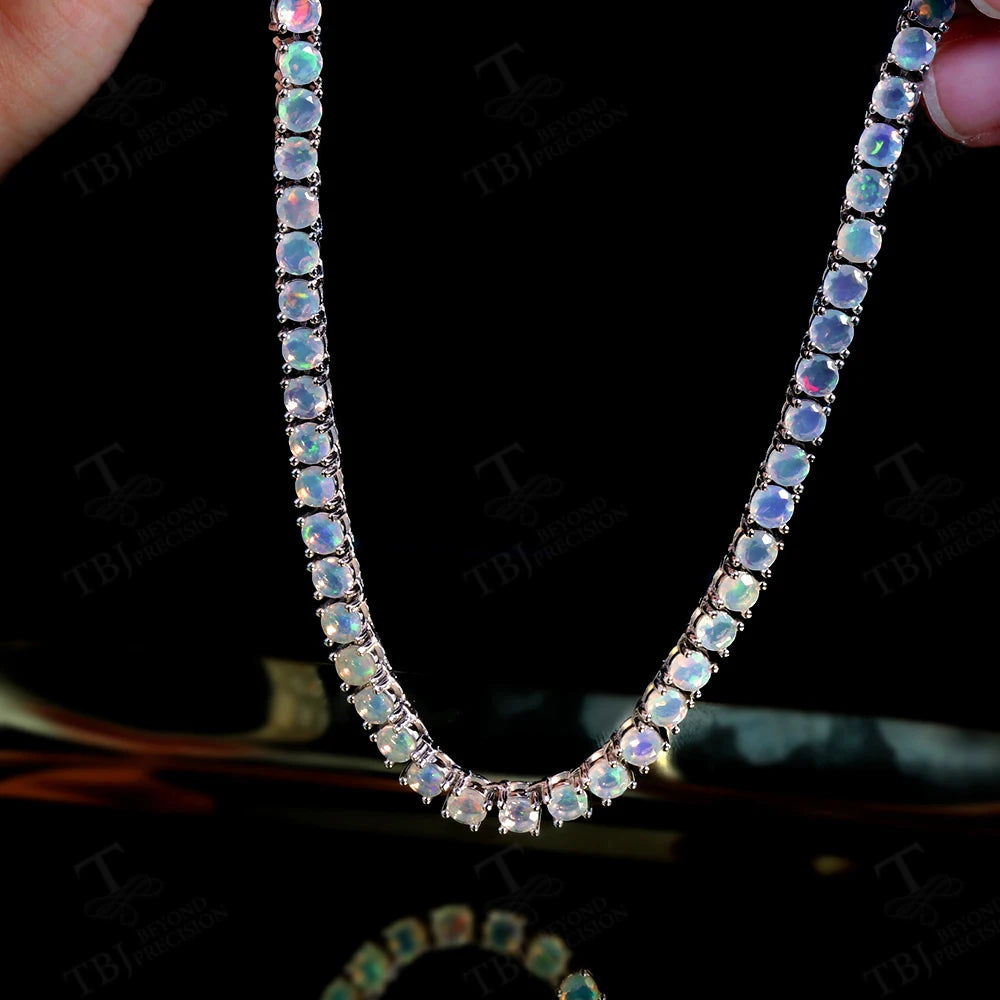 A close-up of a rose gold tennis necklace with iridescent blue and green opal-like round stones, held by fingers against a black background with a faint reflection below.