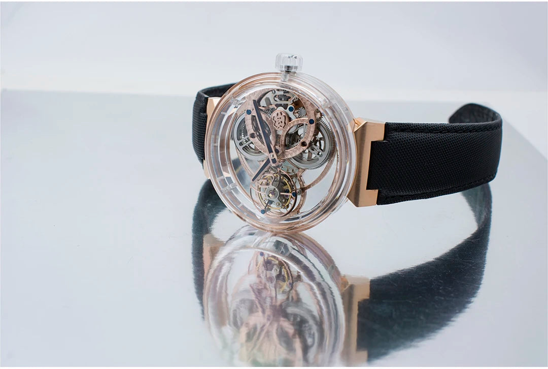 A luxury skeleton watch with a transparent circular case showing a rose gold and silver mechanical movement, paired with a black textured strap, resting on a reflective surface.
