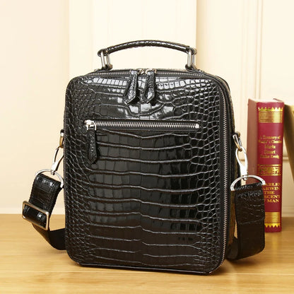 A black crocodile-embossed leather crossbody bag with a top handle, front zip pocket, and detachable shoulder strap, displayed on a wooden surface with books in the background.