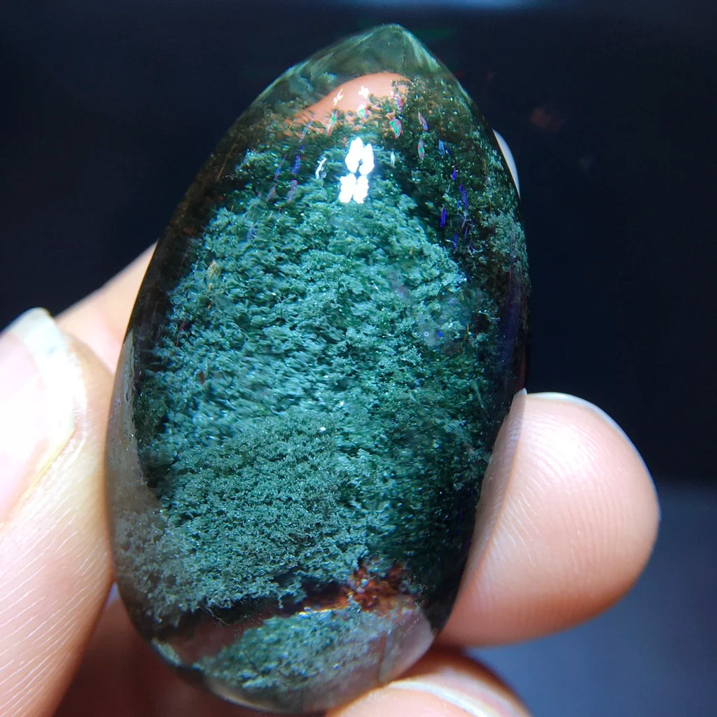 A hand holding a polished green teardrop-shaped gemstone or crystal against a dark background.