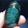 A hand holding a polished green teardrop-shaped gemstone or crystal against a dark background.