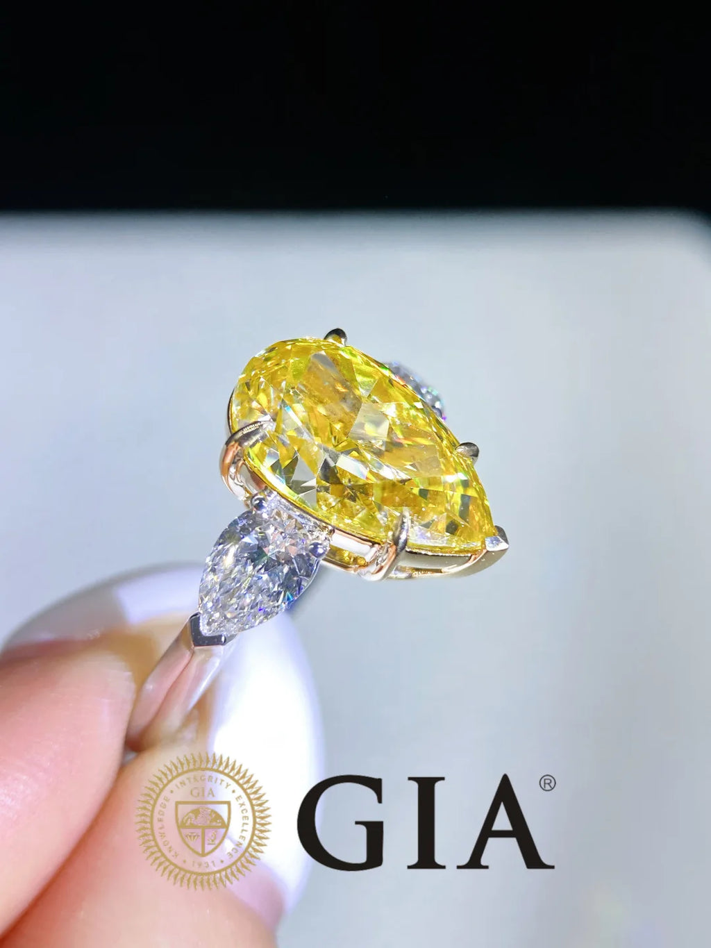 A yellow marquise-cut diamond ring held between fingers, featuring a yellow center stone and a marquise-cut diamond accent on a gold-toned band, with a GIA logo watermark at the bottom.