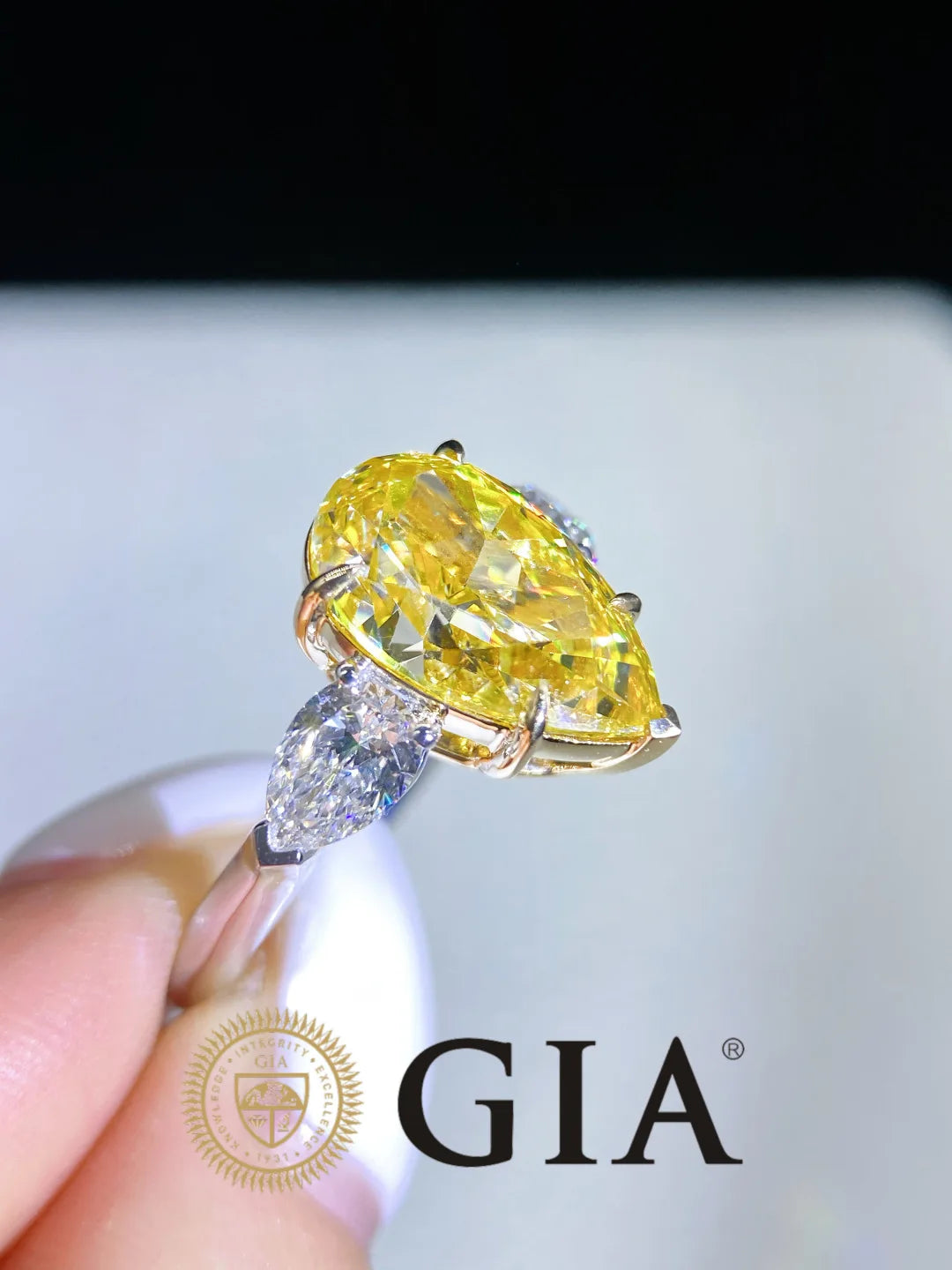 A yellow marquise-cut diamond ring held between fingers, featuring a yellow center stone and a marquise-cut diamond accent on a gold-toned band, with a GIA logo watermark at the bottom.