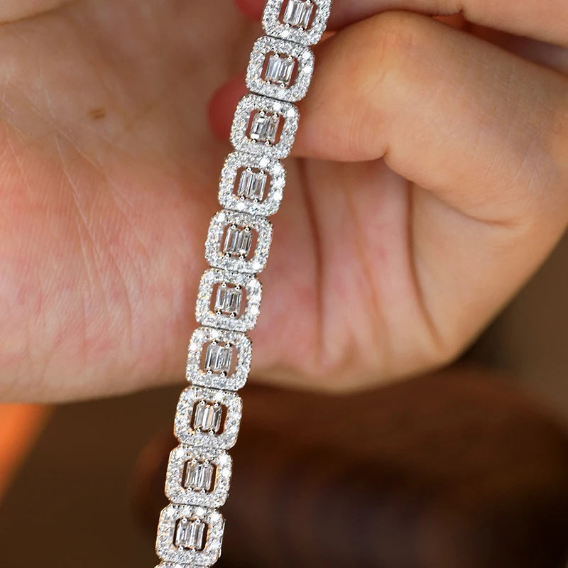 A close-up of a diamond-encrusted tennis bracelet held between fingers. The bracelet features rectangular baguette-cut diamonds set in a silver-toned metal, surrounded by smaller round diamonds.