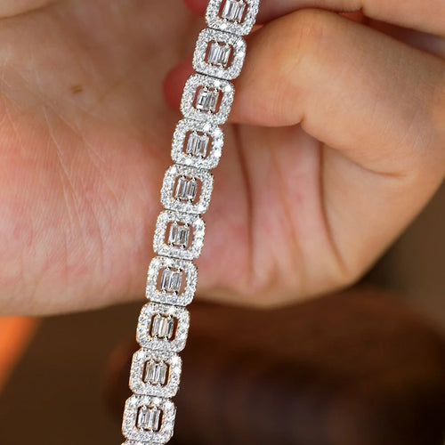 A close-up of a diamond-encrusted tennis bracelet held between fingers. The bracelet features rectangular baguette-cut diamonds set in a silver-toned metal, surrounded by smaller round diamonds.