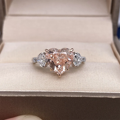 A heart-shaped diamond ring with pear-cut side diamonds set in a rose gold band, resting on a beige velvet-lined jewelry box.