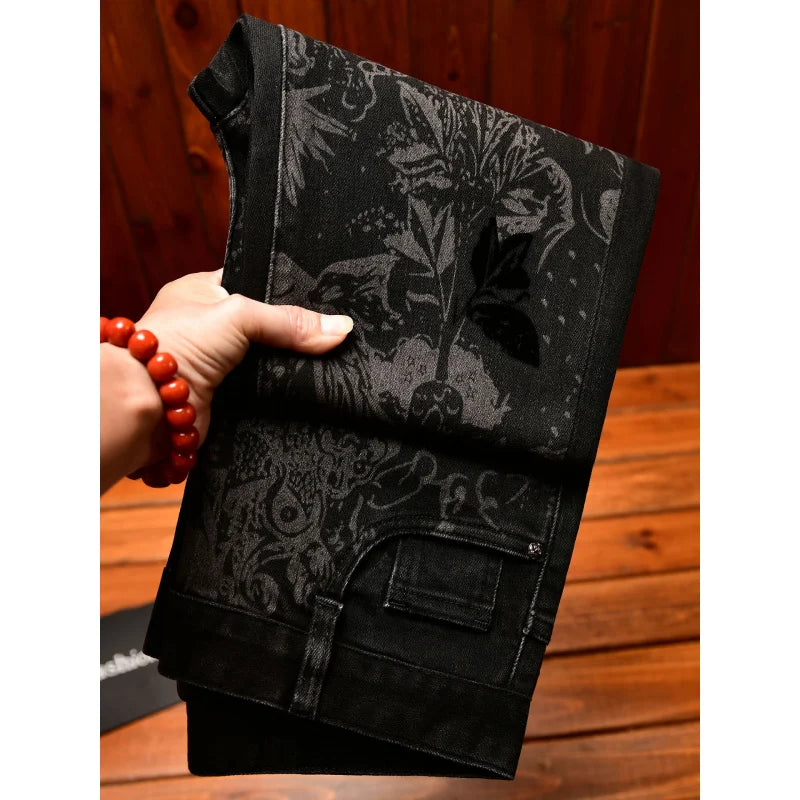 A hand holding a pair of black patterned pants or jeans with a dark botanical and ornamental print, set against a wooden background.