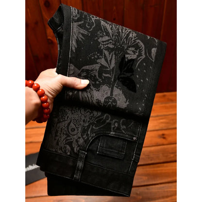 A hand holding a pair of black patterned pants or jeans with a dark botanical and ornamental print, set against a wooden background.