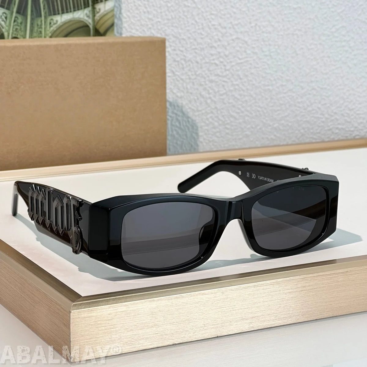 Black square sunglasses with dark lenses and a sculptural geometric temple design, displayed on a light wood tray against a textured white wall.