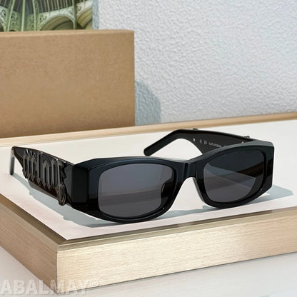 Black square sunglasses with dark lenses and a sculptural geometric temple design, displayed on a light wood tray against a textured white wall.