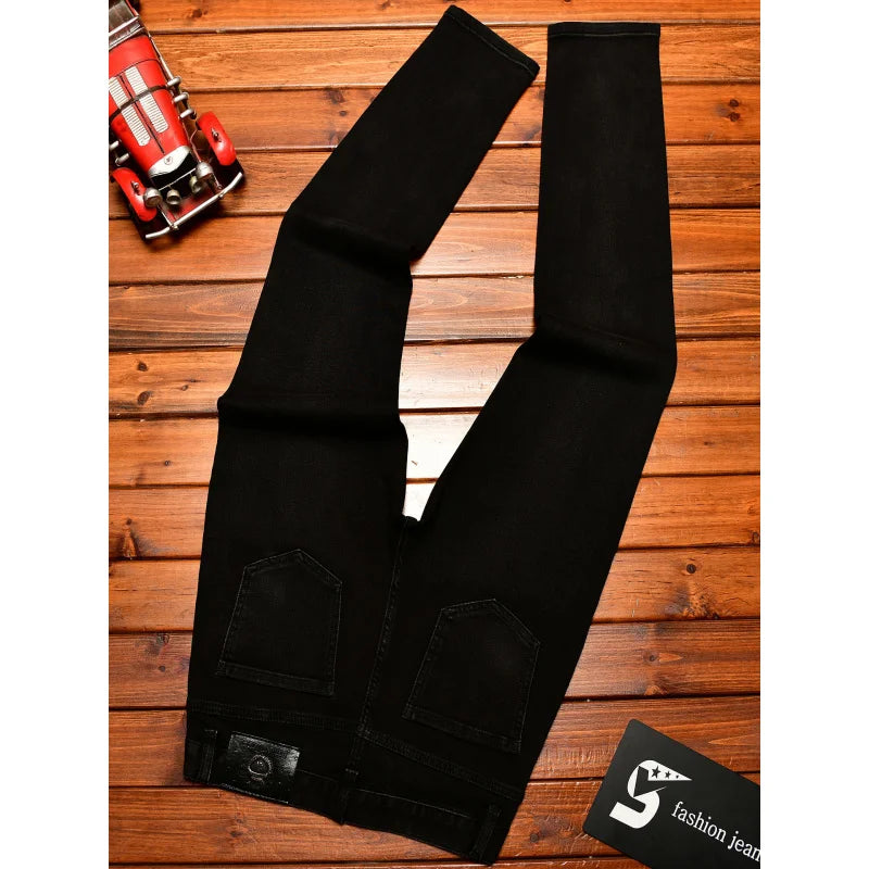 Black denim jeans laid flat on a wooden plank surface, with a small red toy car in the top-left corner and a black card with a white logo in the bottom-right corner.