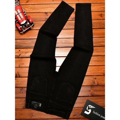 Black denim jeans laid flat on a wooden plank surface, with a small red toy car in the top-left corner and a black card with a white logo in the bottom-right corner.
