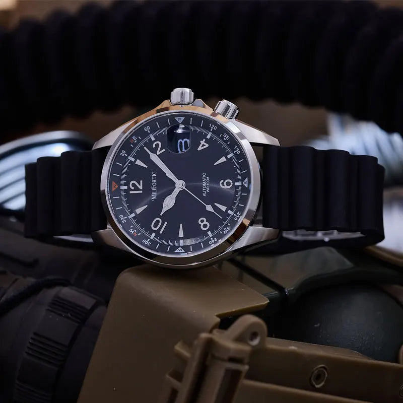 A wristwatch with a black strap, silver-toned metal case, and a dark blue dial featuring white Arabic numerals and a date window, photographed close up against a blurred tactical-style background.