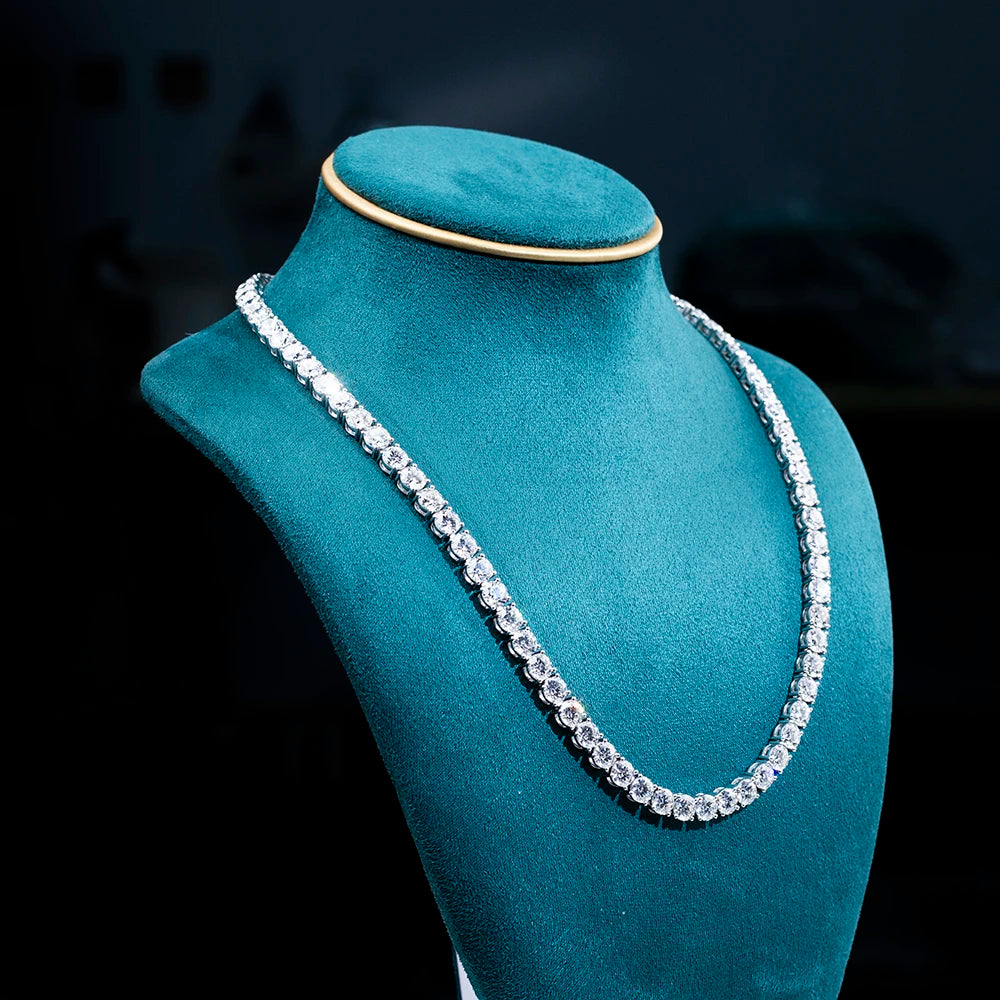 A sparkling silver-toned tennis necklace with clear stones, displayed on a teal velvet jewelry bust against a dark background.